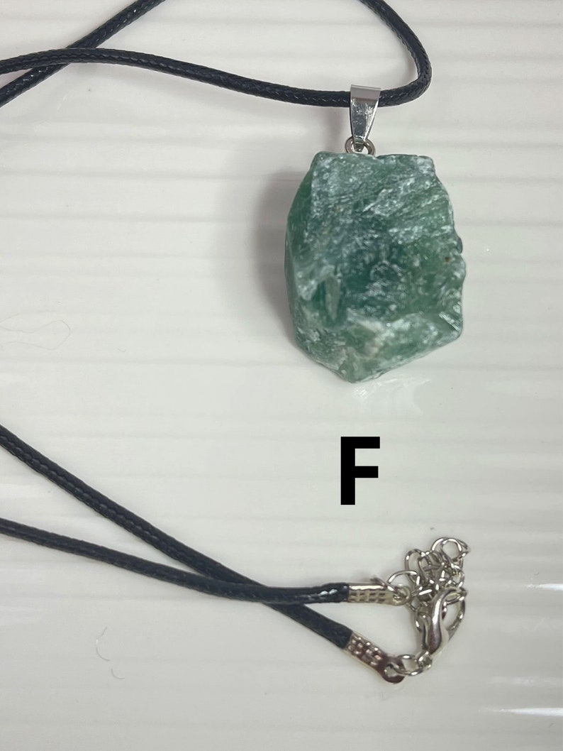 May include: A green stone pendant on a black cord necklace. The pendant is a rough, irregular shape with a silver bail. The necklace has a silver clasp. The letter "F" is printed on a white surface below the necklace.