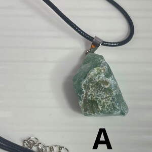 May include: A green stone pendant on a black cord necklace. The stone is a rough, irregular shape and has a light green color with white flecks. The pendant is attached to the cord with a silver bail. The necklace has a silver clasp.