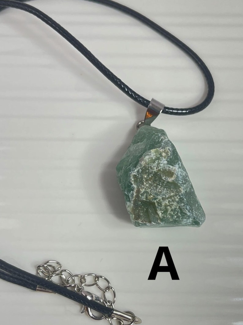 May include: A green stone pendant on a black cord necklace. The stone is a rough, irregular shape and has a light green color with white flecks. The pendant is attached to the cord with a silver bail. The necklace has a silver clasp.