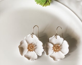 May include: A pair of white flower earrings with gold-colored centers and ear wires. The earrings are shaped like open blossoms, with detailed petal textures. The earrings are displayed on a white plate, with green flowers in the background.
