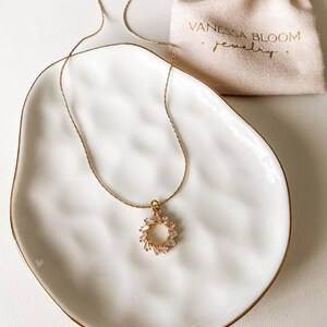 Pu&ograve; includere: Una collana con catena dorata e un delicato ciondolo a forma di cerchio di piccoli cristalli trasparenti. La collana &egrave; esposta su un piatto in ceramica bianca con un bordo dorato. Il piatto &egrave; appoggiato su una superficie bianca. Il testo "Vanessa Bloom Jewelry" &egrave; visibile su una piccola borsa beige.