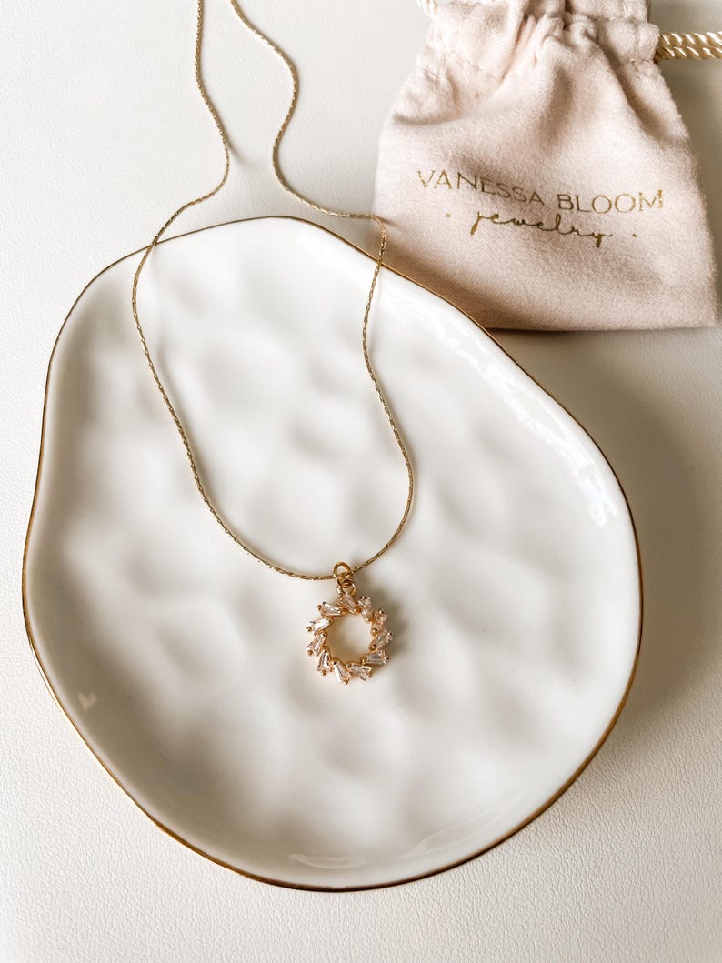 Pu&ograve; includere: Una collana con catena dorata e un delicato ciondolo a forma di cerchio di piccoli cristalli trasparenti. La collana &egrave; esposta su un piatto in ceramica bianca con un bordo dorato. Il piatto &egrave; appoggiato su una superficie bianca. Il testo "Vanessa Bloom Jewelry" &egrave; visibile su una piccola borsa beige.