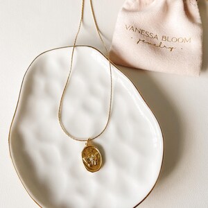 Pu&ograve; includere: Una collana con catena dorata con un ciondolo ovale dorato con un design a farfalla. La collana &egrave; esposta su un piatto in ceramica bianca con un bordo dorato. La collana &egrave; confezionata in una piccola borsa bianca con il testo "Vanessa Bloom Gioielli".