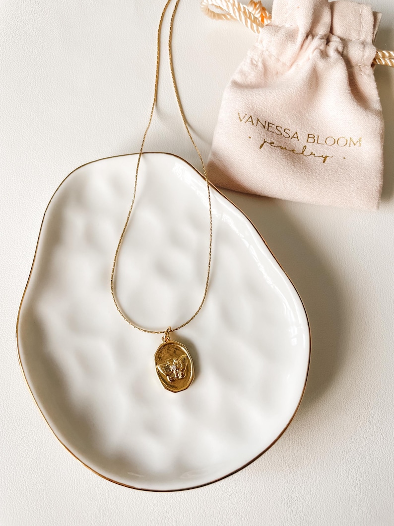 Pu&ograve; includere: Una collana con catena dorata con un ciondolo ovale dorato con un design a farfalla. La collana &egrave; esposta su un piatto in ceramica bianca con un bordo dorato. La collana &egrave; confezionata in una piccola borsa bianca con il testo "Vanessa Bloom Gioielli".