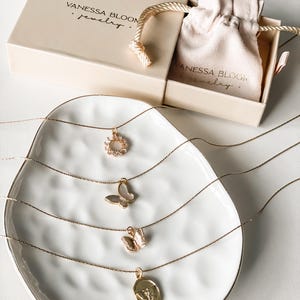 Pu&ograve; includere: Quattro collane d'oro con ciondoli diversi su un piatto bianco. I ciondoli includono un fiore, una farfalla, una farfalla pi&ugrave; piccola e un ciondolo rotondo con un design. Le collane sono esposte su un piatto bianco con una superficie testurizzata. Il testo "Vanessa Bloom Jewelry" &egrave; visibile sulla scatola sullo sfondo.