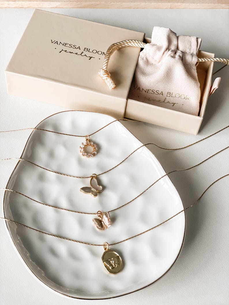 Pu&ograve; includere: Quattro collane d'oro con ciondoli diversi su un piatto bianco. I ciondoli includono un fiore, una farfalla, una farfalla pi&ugrave; piccola e un ciondolo rotondo con un design. Le collane sono esposte su un piatto bianco con una superficie testurizzata. Il testo "Vanessa Bloom Jewelry" &egrave; visibile sulla scatola sullo sfondo.