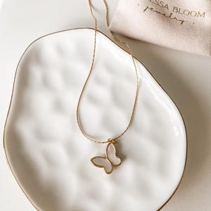Pu&ograve; includere: Una collana a catena dorata con un ciondolo a forma di farfalla bianca. La collana &egrave; su un piatto di ceramica bianca. Il piatto ha un bordo dorato. La collana &egrave; in una piccola borsa bianca con il testo "Vanessa Bloom Jewelry".