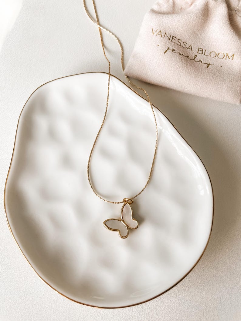 Pu&ograve; includere: Una collana a catena dorata con un ciondolo a forma di farfalla bianca. La collana &egrave; su un piatto di ceramica bianca. Il piatto ha un bordo dorato. La collana &egrave; in una piccola borsa bianca con il testo "Vanessa Bloom Jewelry".