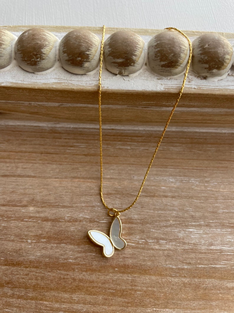 Pu&ograve; includere: Una collana con catena dorata e un ciondolo a forma di farfalla bianca. La farfalla &egrave; realizzata in madreperla e ha un contorno dorato.