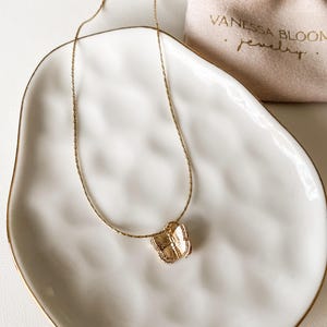 Pu&ograve; includere: Una collana con catena dorata e un delicato ciondolo a forma di farfalla. La collana &egrave; esposta su un piatto in ceramica bianca con un bordo dorato. Il piatto &egrave; appoggiato su una superficie bianca. La collana &egrave; confezionata in una piccola busta bianca con il testo "Vanessa Bloom Jewelry" stampato in oro.