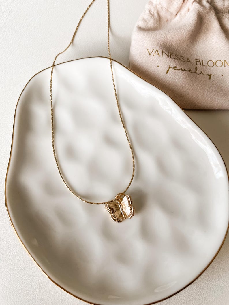 Pu&ograve; includere: Una collana con catena dorata e un delicato ciondolo a forma di farfalla. La collana &egrave; esposta su un piatto in ceramica bianca con un bordo dorato. Il piatto &egrave; appoggiato su una superficie bianca. La collana &egrave; confezionata in una piccola busta bianca con il testo "Vanessa Bloom Jewelry" stampato in oro.