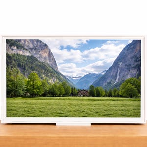 May include: A white framed digital display shows a scenic landscape. The image features a green meadow, a small wooden cabin, and towering mountains under a blue sky with fluffy clouds. The display sits on a wooden surface.