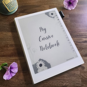 May include: A white digital notebook displays the text "My Cursive Notebook" in cursive script. Floral illustrations and a daisy accent the corners. A black stylus rests on the side. A purple flower and a green jar are also visible on the wooden surface.