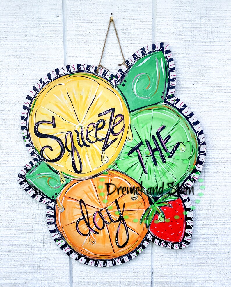May include: A wooden door hanger with a painted design of lemons, limes, and an orange. The text "Squeeze the day" is written in black paint. A red strawberry is painted in the bottom right corner.