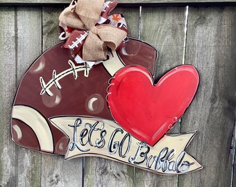 May include: A brown and white football with a red heart and a banner that says "Let's Go Buffalo". The football has a brown burlap bow on top.