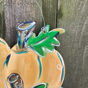 May include: A wooden pumpkin decoration with a wire hanger. The pumpkin is painted orange with green and white accents, and a green leaf. The background is a weathered wooden fence.