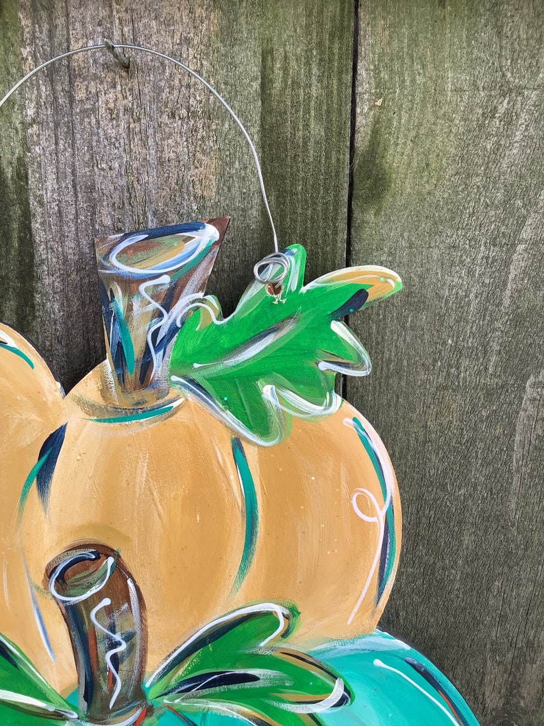 May include: A wooden pumpkin decoration with a wire hanger. The pumpkin is painted orange with green and white accents, and a green leaf. The background is a weathered wooden fence.