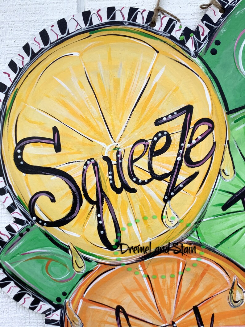 May include: A painted wooden door hanger with three citrus fruits, a lemon, an orange, and a lime. The lemon has the word "Squeeze" written in black with white dots on it. The background is black and white stripes with a red and white pattern.