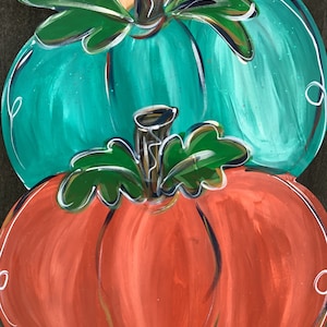 May include: A wooden, stacked pumpkin decoration. The bottom pumpkin is orange, the middle is teal, and the top is tan. Each pumpkin has green leaves and a brown stem. White swirls accent the edges of each pumpkin.