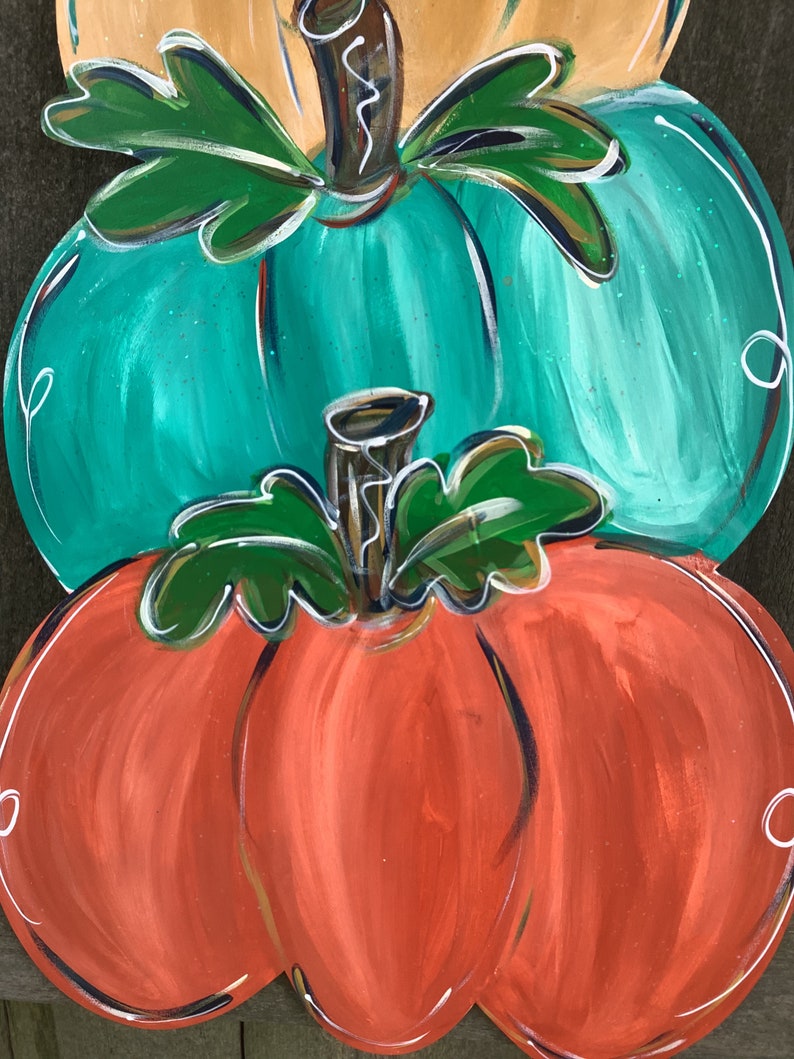 May include: A wooden, stacked pumpkin decoration. The bottom pumpkin is orange, the middle is teal, and the top is tan. Each pumpkin has green leaves and a brown stem. White swirls accent the edges of each pumpkin.