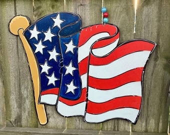 May include: A wooden American flag door hanger with red, white, and blue stripes and white stars on a blue field. The flag is attached to a wooden pole with a gold tip.