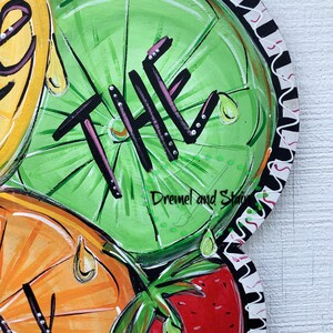 May include: A wooden doormat with a painted design of fruit slices, including a lime with the word "THE" painted on it, an orange, and a strawberry. The doormat has a black and white scalloped border.