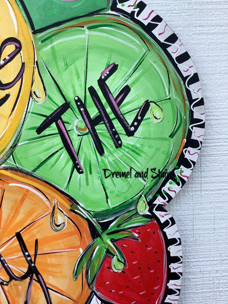 May include: A wooden doormat with a painted design of fruit slices, including a lime with the word "THE" painted on it, an orange, and a strawberry. The doormat has a black and white scalloped border.