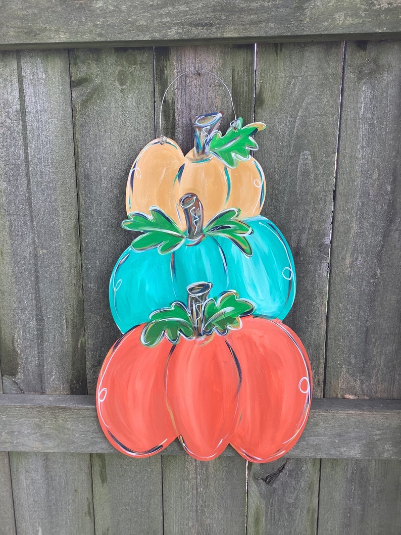 May include: A wooden pumpkin decoration with three stacked pumpkins in orange, teal, and yellow. Each pumpkin has a green stem and leaf detail, outlined in black. A wire loop is attached for hanging. The background is a wooden fence.