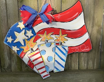 May include: A wooden door hanger decorated with a red, white, and blue American flag with three sparklers. The sparklers are painted with red, white, and blue stripes and polka dots.