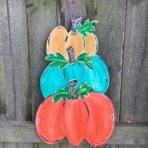 May include: A wooden pumpkin decoration with three stacked pumpkins in orange, teal, and yellow. Each pumpkin has a green stem and leaf detail, with white and black accents. A wire loop is attached for hanging.
