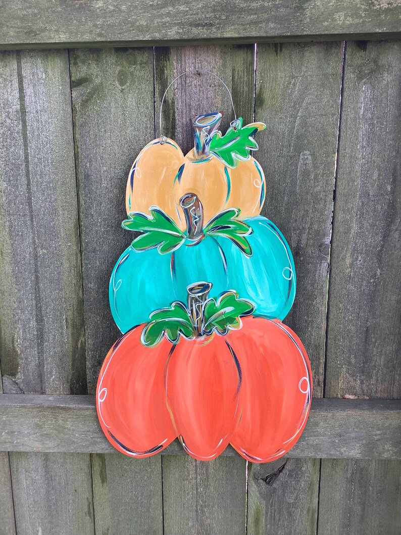May include: A wooden pumpkin decoration with three stacked pumpkins in orange, teal, and yellow. Each pumpkin has a green stem and leaf detail, with white and black accents. A wire loop is attached for hanging.