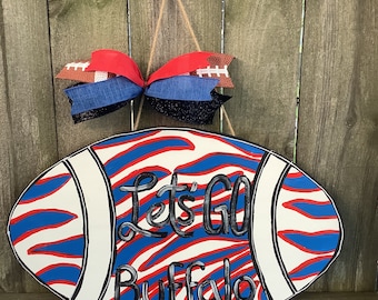 May include: A football-shaped door hanger with a blue and red zebra stripe pattern. The words "Let's Go Buffalo" are written in black on the front of the hanger. A red, white, and blue bow with football-shaped accents is attached to the top of the hanger.