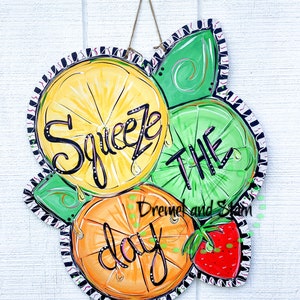 May include: A wooden door hanger with a painted design of lemons, limes, and an orange. The text "Squeeze the day" is written in black paint. A strawberry is painted in the bottom right corner.