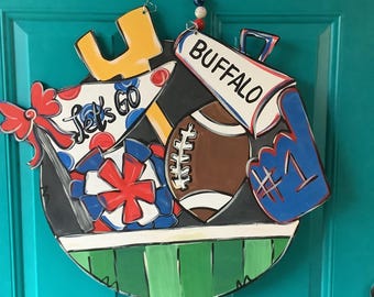 May include: A round, wooden door hanger with a football theme. It features a football, a foam finger with the number 1, a megaphone with "BUFFALO" on it, and the words "Let's Go". The color scheme includes red, white, blue, yellow, green, and brown.