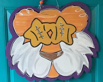 May include: A decorative wooden door hanger in the shape of a tiger's face. The tiger has an orange and purple outline, with white fur, a brown nose, and a yellow bow tie. The word "GO" is written on the bow tie.