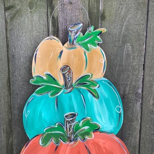 May include: A wooden pumpkin decoration with three stacked pumpkins in orange, teal, and yellow. Each pumpkin has a green leaf and a silver stem. The decoration is hanging from a silver wire and is set against a wooden fence.