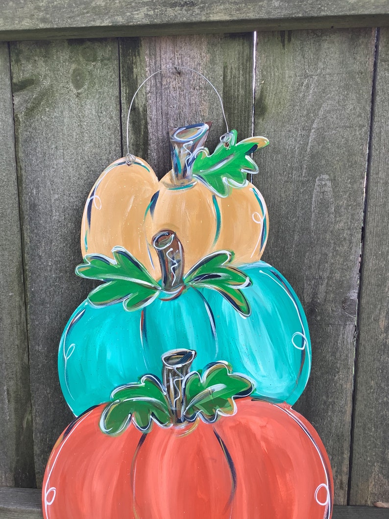 May include: A wooden pumpkin decoration with three stacked pumpkins in orange, teal, and yellow. Each pumpkin has a green leaf and a silver stem. The decoration is hanging from a silver wire and is set against a wooden fence.