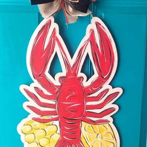 May include: A red lobster with yellow corn and lemons painted on a wooden door hanger. The lobster is painted in a realistic style with black outlines. The hanger is decorated with a black and white checkered bow.