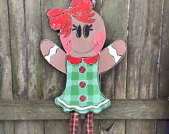 May include: A gingerbread girl Christmas decoration with a red bow, green and white plaid dress, and red and white plaid leggings. The gingerbread girl is hanging from a wooden fence.