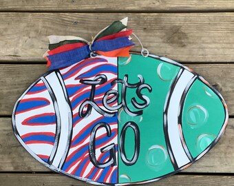 May include: A green and white football-shaped door hanger with red, white, and blue stripes on one side. The words "Let's Go" are written in black on the door hanger.