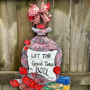 May include: A wooden sign with a red and white checkered bag, a red lobster, corn on the cob, lemons, and a blue and yellow can. The sign says "Let the Good Times Boil".