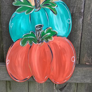 May include: A stacked pumpkin decoration with three pumpkins in orange, teal, and tan. Each pumpkin has a green stem and leaves, with white accents outlining the edges. The pumpkins are painted with a glossy finish.