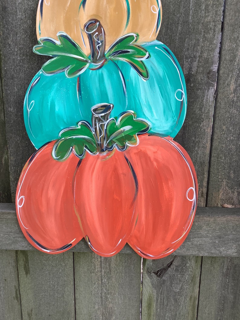 May include: A stacked pumpkin decoration with three pumpkins in orange, teal, and tan. Each pumpkin has a green stem and leaves, with white accents outlining the edges. The pumpkins are painted with a glossy finish.