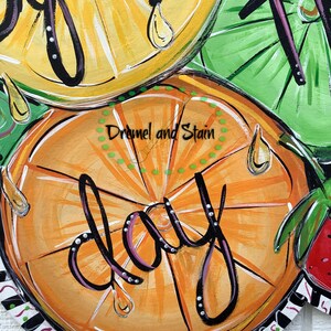 May include: A painted wooden door hanger with a citrus theme. The hanger features a large orange slice with the word "day" written in black script. The orange slice is surrounded by lemons and lime slices, all with painted details of leaves and dripping juice.