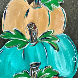 May include: A stacked, hand-painted wooden pumpkin decoration. The top pumpkin is yellow, the middle is teal, and the bottom is orange. Each pumpkin has a brown stem and green leaves. White accents and a wire hanger complete the design.