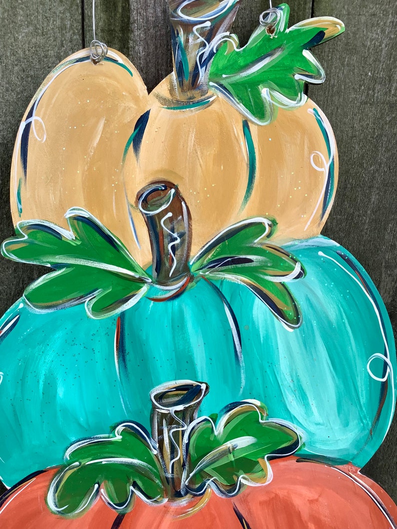 May include: A stacked, hand-painted wooden pumpkin decoration. The top pumpkin is yellow, the middle is teal, and the bottom is orange. Each pumpkin has a brown stem and green leaves. White accents and a wire hanger complete the design.