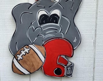 May include: A gray elephant-shaped wooden door hanger with a football and a red helmet. The elephant has black and white details, and the football is brown with white stripes. A wire and beads are attached for hanging.