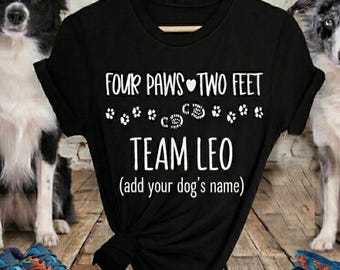 May include: A black t-shirt featuring white text that reads Four Paws Two Feet Team Leo (add your dog's name) with a series of small paw print graphics and a heart symbol centered on the front of the garment, displayed on a wooden hanger.