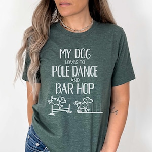 May include: A green t-shirt with the text "My dog loves to pole dance and bar hop" in white. The shirt features a cartoon illustration of a dog jumping over a hurdle and another dog running through a set of poles.