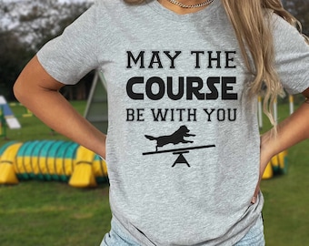 May include: A light gray t-shirt with the text "MAY THE COURSE BE WITH YOU" and a dog silhouette on a seesaw. The shirt is made of a soft material and is perfect for dog lovers.